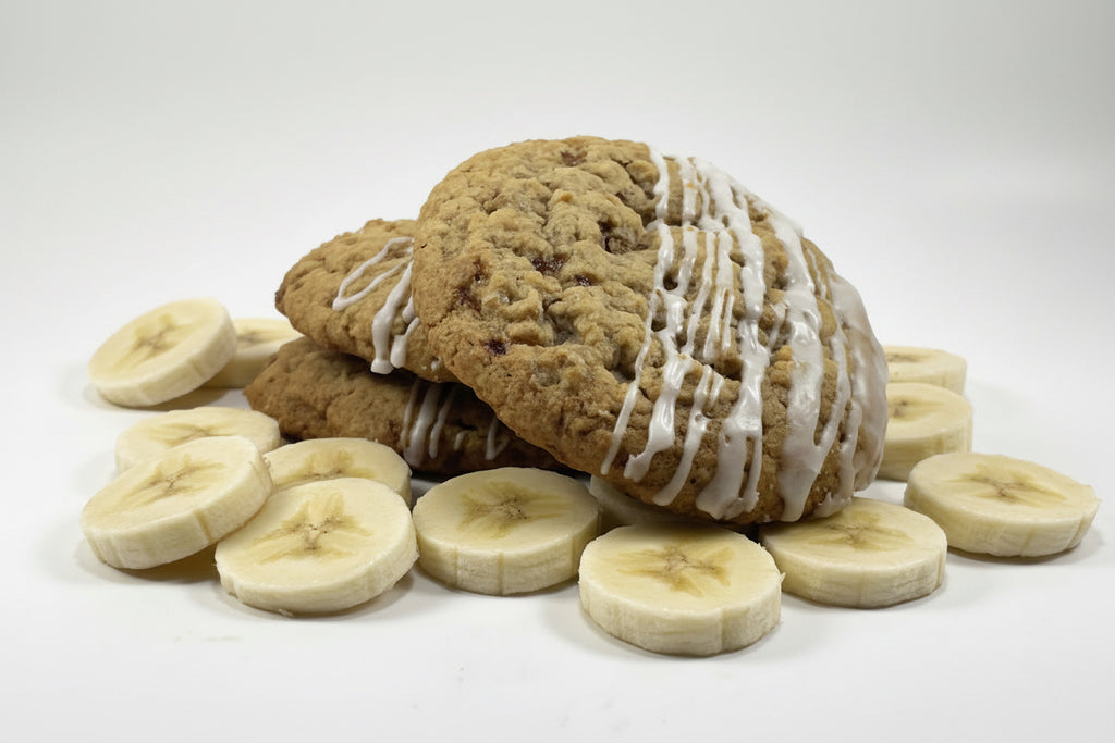 Side view of three thick, golden-brown cookies stacked slightly offset, with a soft, crumbly texture and white icing drizzled over the tops, photographed on a clean white background with banana slices around the cookie.